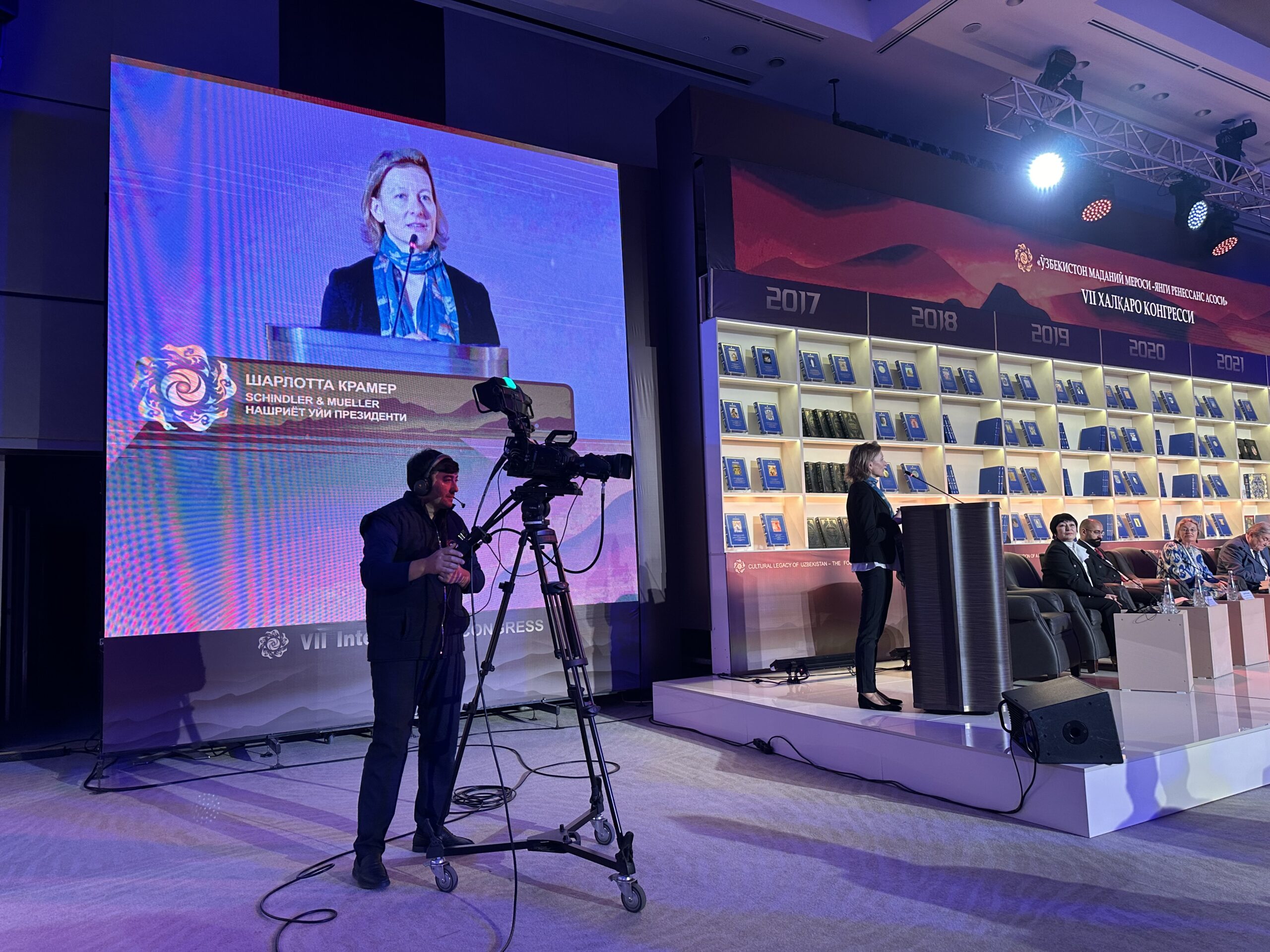 Eindrücke aus Samarkand, Usbekistan: Unsere Verlegerin Charlotte Kramer hinter dem Rednerpult auf der Bühne mit Kamera und Bildeinblendung auf deiner grossen Leinwand